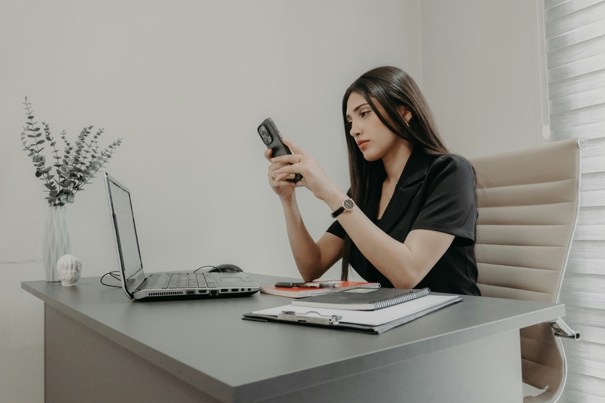 Professional woman at a modern desk with laptop and notebooks, checking her phone while working in a bright home office
